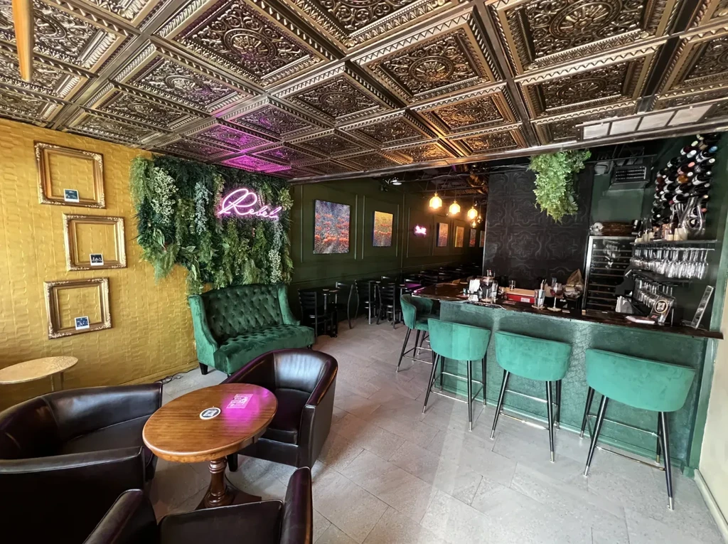 Empty bar with gold ceiling, green velvet, and 'Rebel' neon sign.