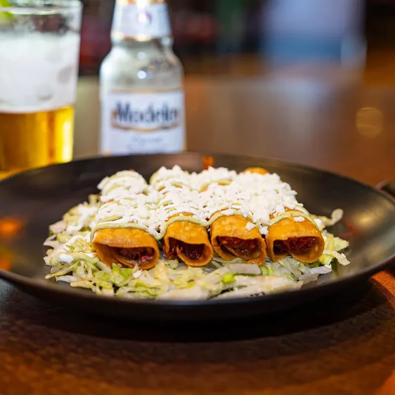 Taquitos topped with cheese, green sauce, lettuce, and Modelo beer.