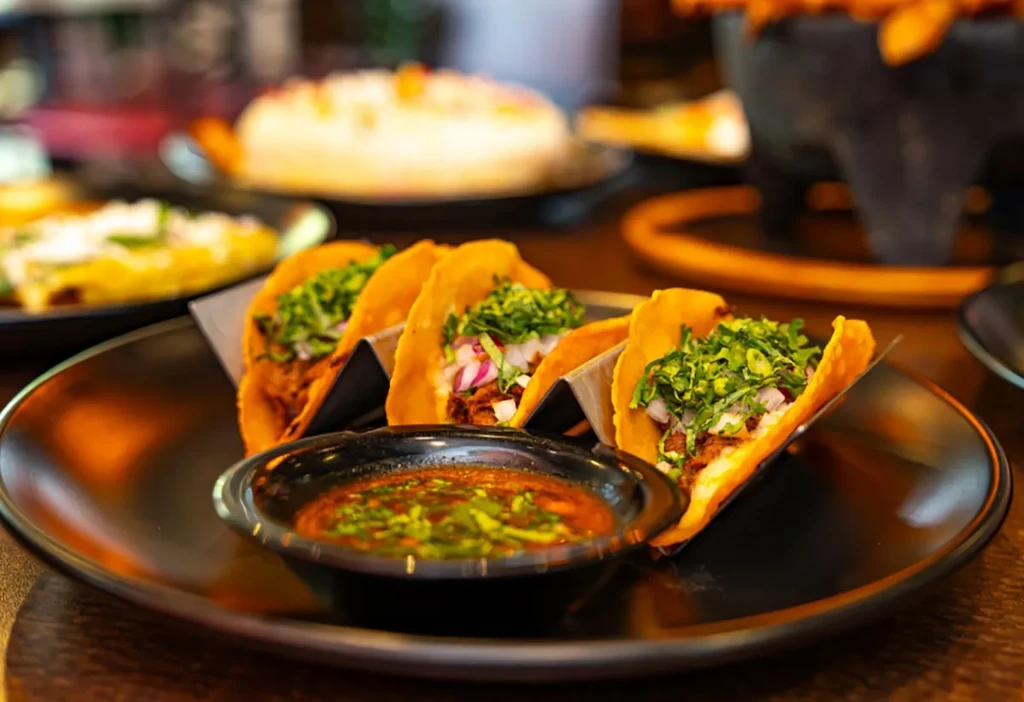 Four golden-brown meat tacos with herbs, onions, and side salsa.