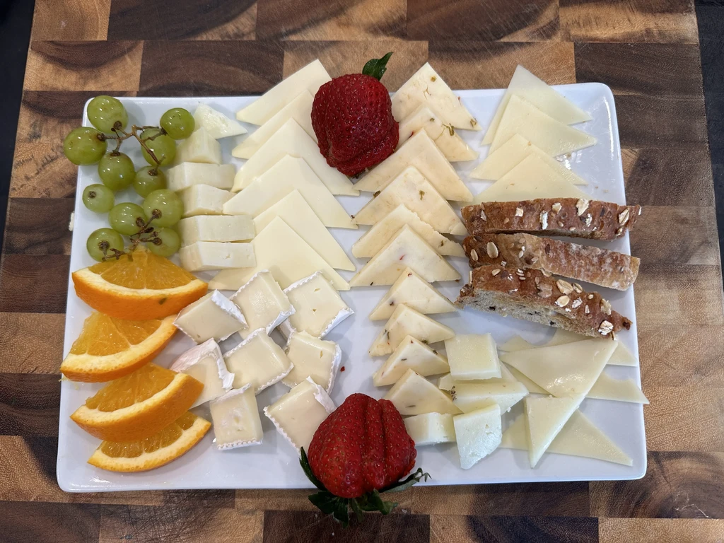 White platter with cheeses, grapes, orange, strawberries, and multi-grain bread.
