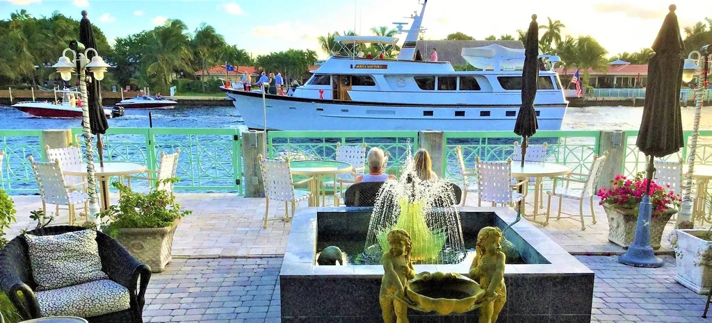 Waterfront dining patio with cherub fountain, yacht, and seated people.