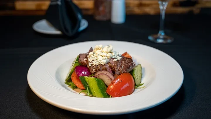 Grilled steak salad with blue cheese, tomatoes, and greens on a white plate.
