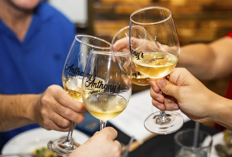 Four hands clinking 'Anthony's Clam House & Grill' wine glasses.