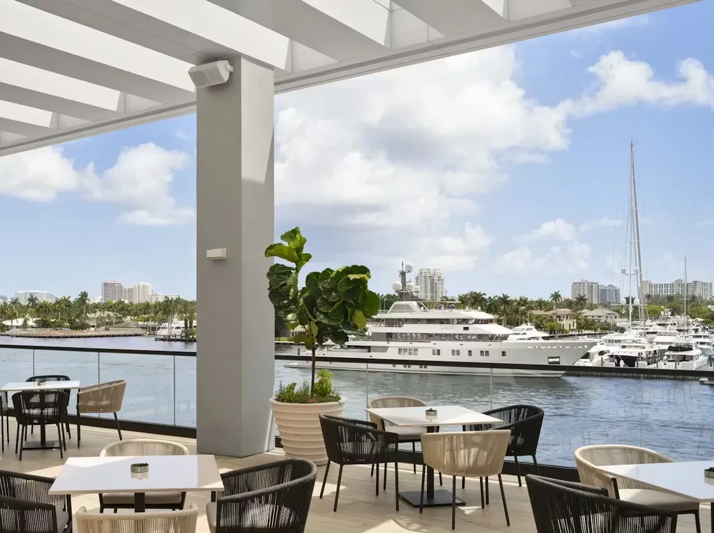 Modern outdoor dining patio overlooks a sunny waterfront filled with yachts.