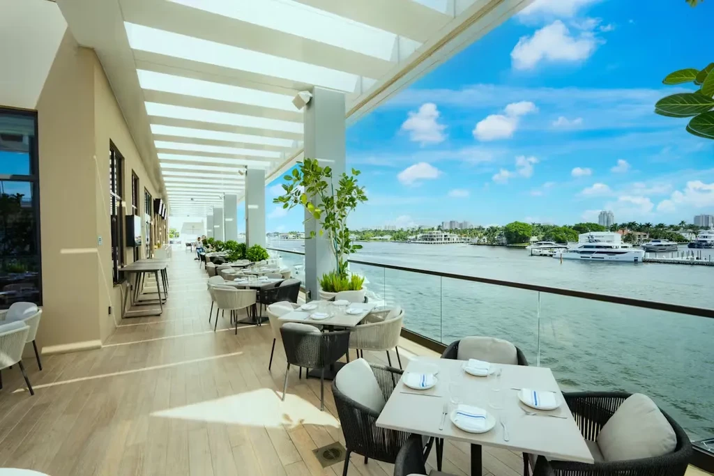 Waterfront dining patio under a pergola, overlooking yachts and cityscape.