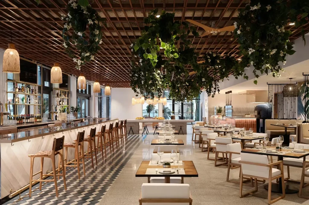 Modern restaurant with long bar, dining tables, and tropical hanging plants.