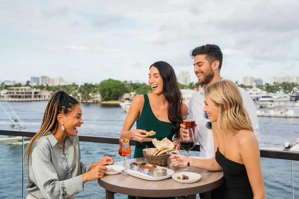 Four friends laugh, enjoying food and drinks on a waterfront balcony.