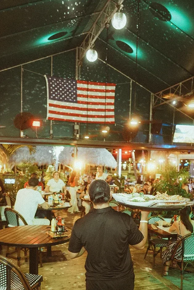 Waiter carries food in a lively tropical outdoor restaurant at night.