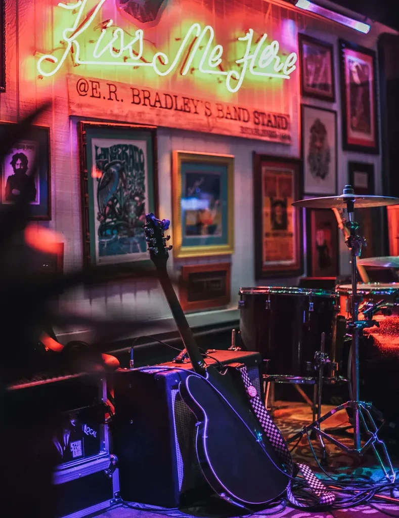 Dimly lit stage with instruments, colored lights, and 'Kiss Me Here' neon sign.
