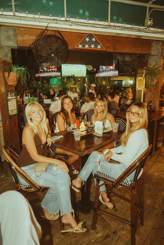 Four young women smile at the camera in a busy sports bar.