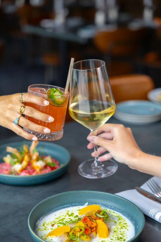 Hands toast cocktail and wine, with colorful seafood dishes at a restaurant.