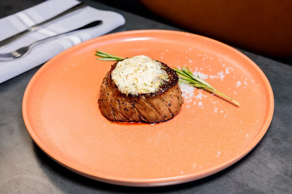 Steak topped with herb butter, rosemary, and salt on a coral plate.