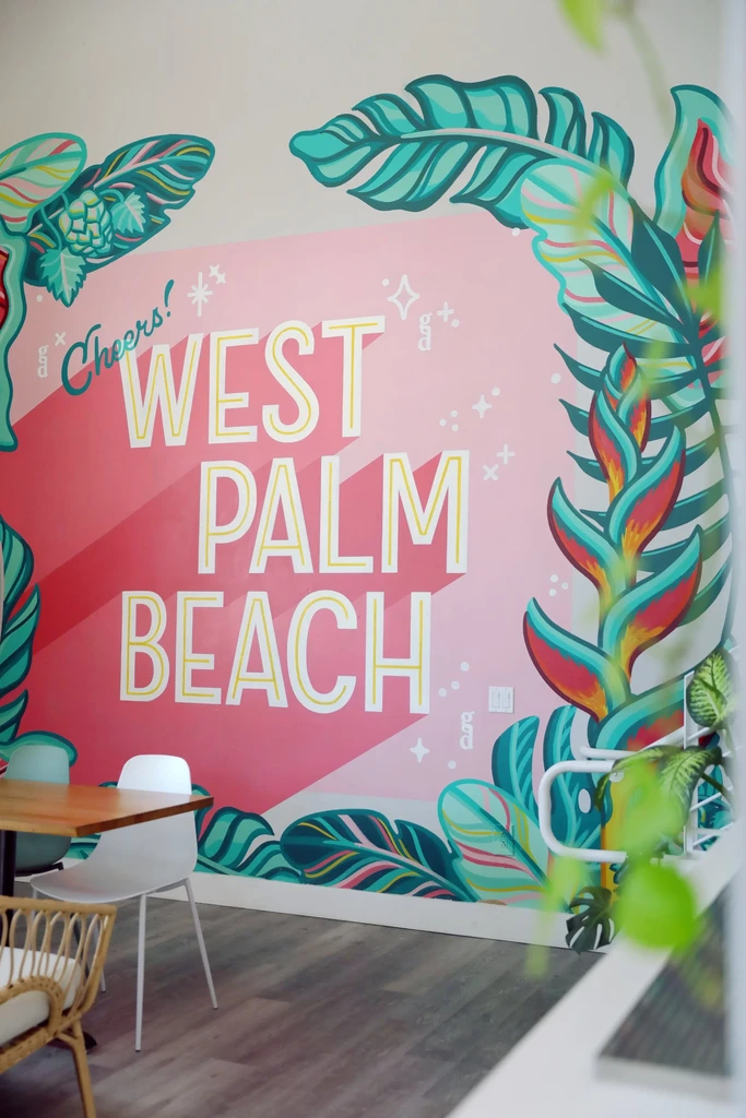 Restaurant interior with tropical mural spelling 'WEST PALM BEACH Cheers!'.