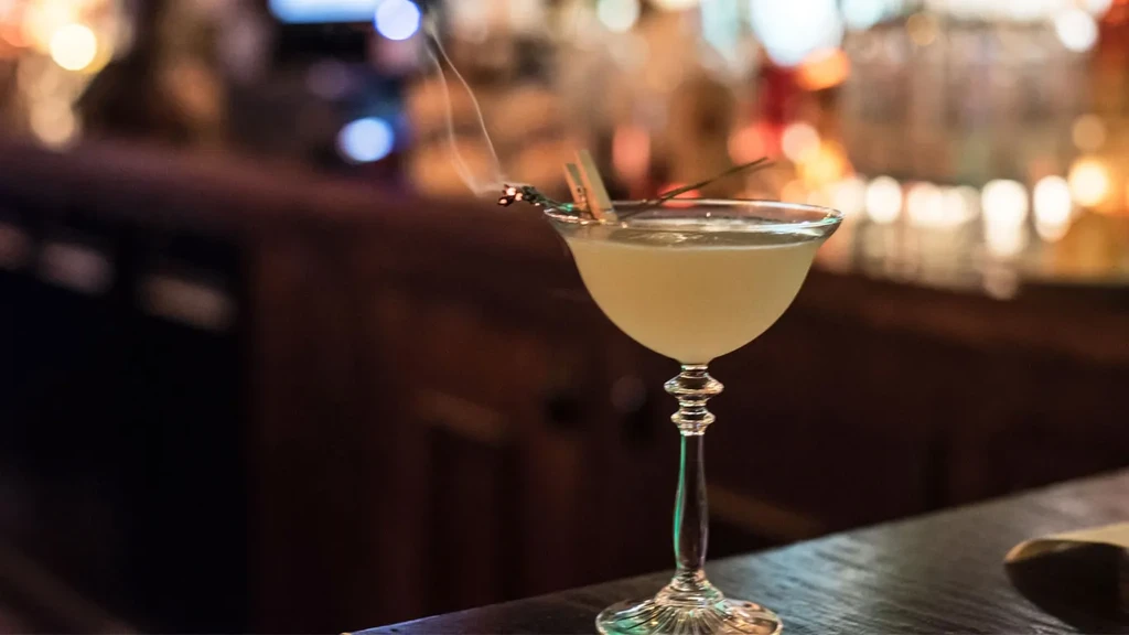 Light cocktail in ornate coupe glass, smoking garnish, blurred bar.
