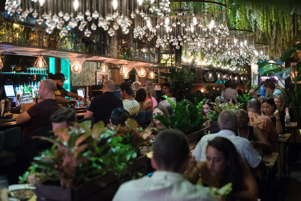 Bustling restaurant interior with stylish lighting, lush foliage, and many patrons.