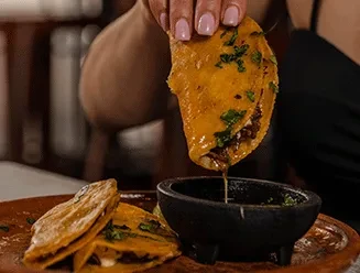 Hand dips golden-brown birria taco into small sauce bowl.