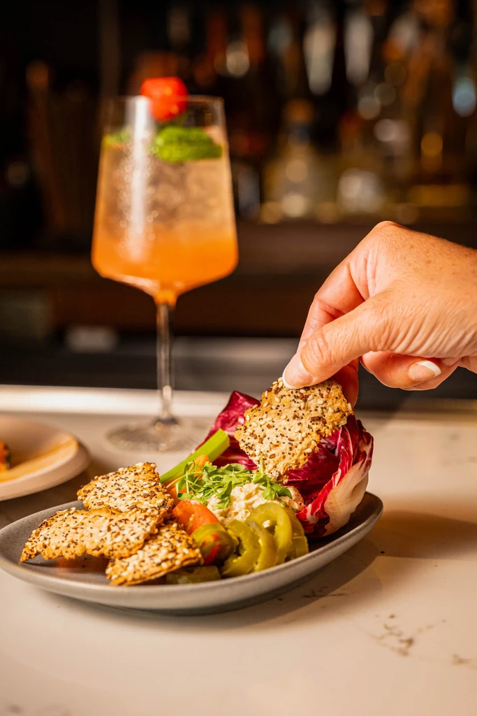Hand takes seeded cracker from dip platter with radicchio and drink.