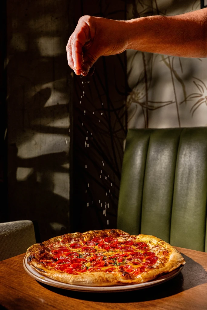 Hand sprinkles seasoning on a freshly baked, cheesy pepperoni pizza.