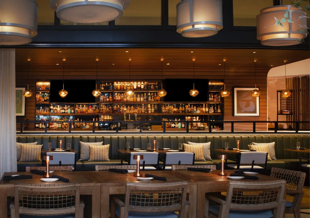Warm-lit modern restaurant interior with communal table, booths, and stocked bar.
