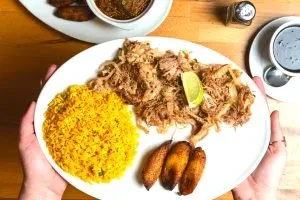 Hands hold a plate with shredded meat, yellow rice, and fried plantains.
