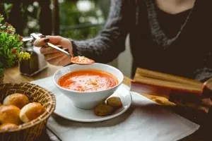 Close-up of person eating creamy orange soup, bread, and reading a book.