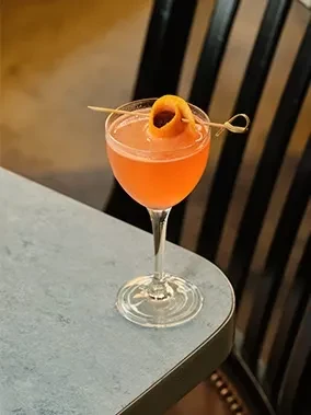 Vibrant coral cocktail in coupe glass with orange peel garnish.