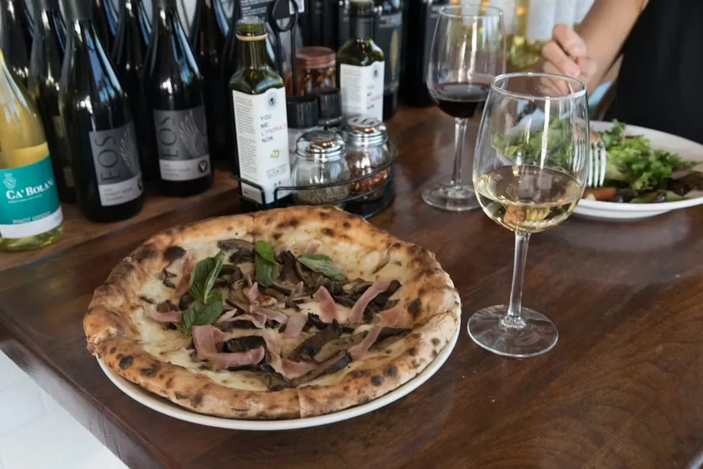Wood-fired pizza, white and red wine, and salad on a dark table.