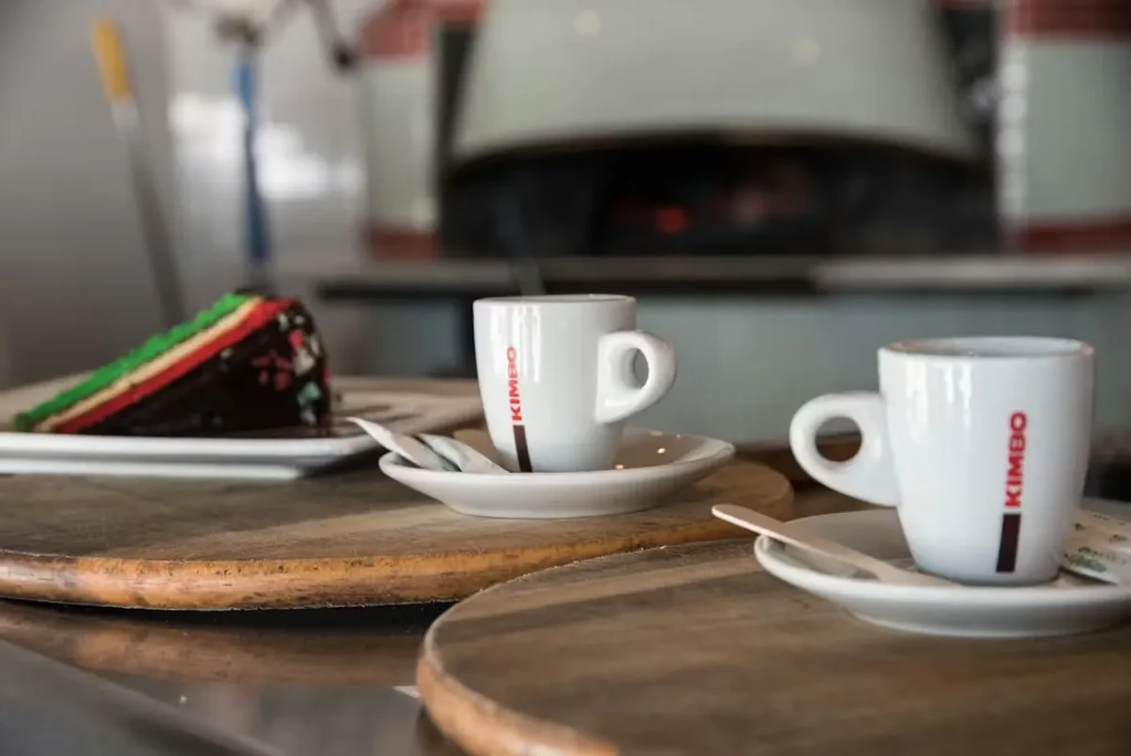 Two Kimbo espressos and layered cake on counter near a pizza oven.