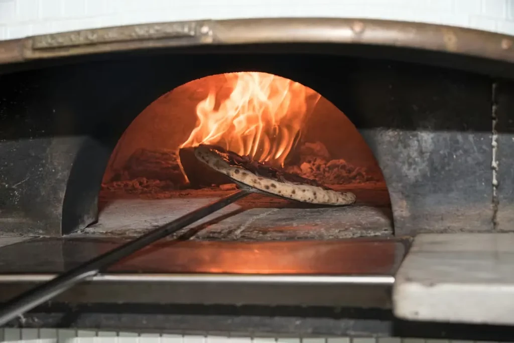 Charred pizza on a peel cooking in a glowing wood-fired oven.