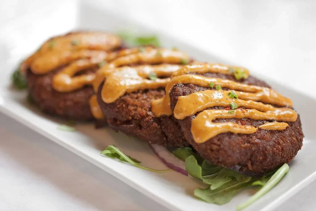 Three seared dark brown patties with orange sauce, herbs, and greens.