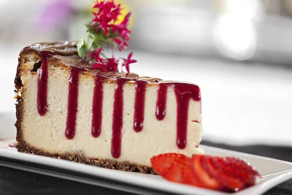 Plated cheesecake slice with berry sauce, flowers, and sliced strawberries.