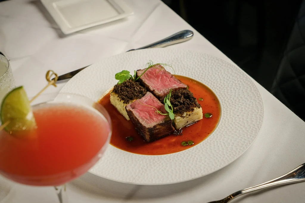Gourmet medium-rare beef, layered potatoes, sauce, microgreens, and a cocktail.