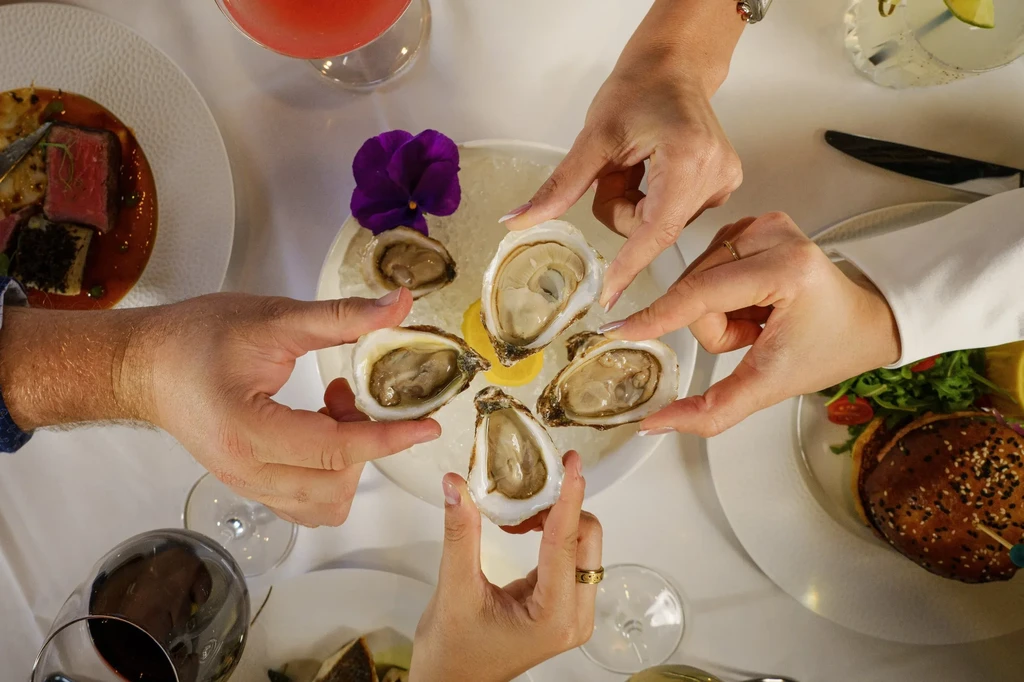 Overhead view of hands sharing oysters, various dishes and drinks.