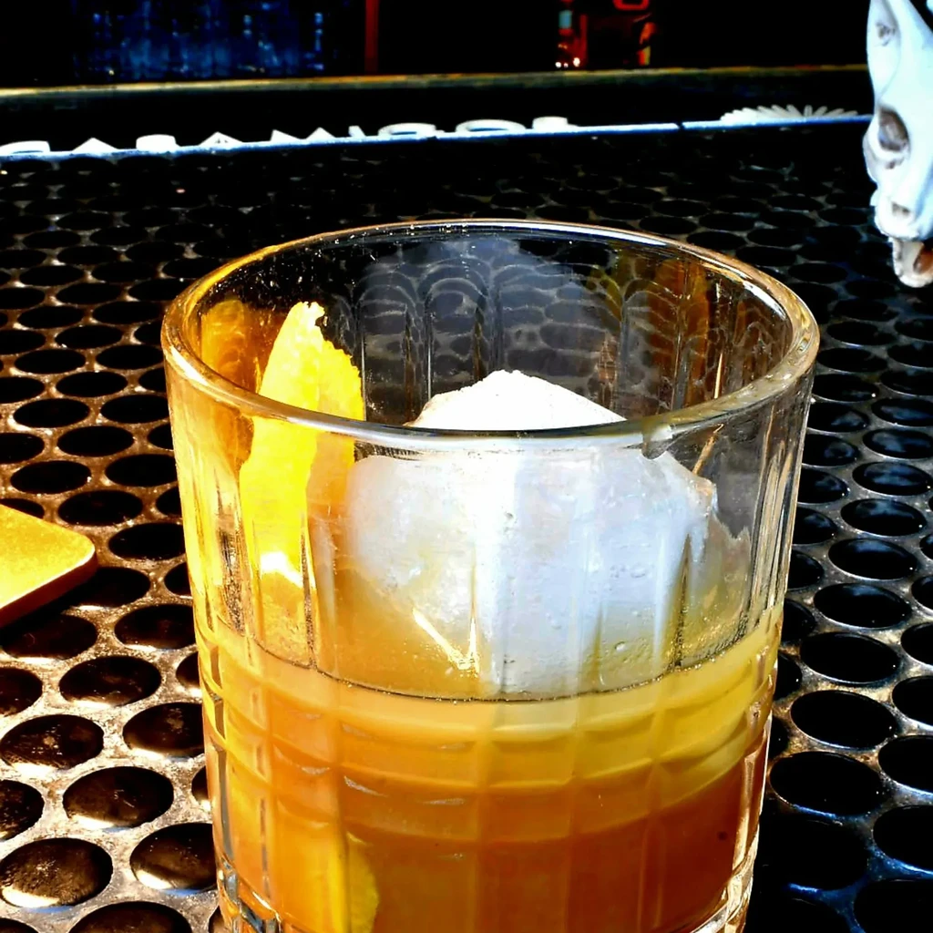 Golden-brown cocktail in textured glass with square ice and citrus peel.