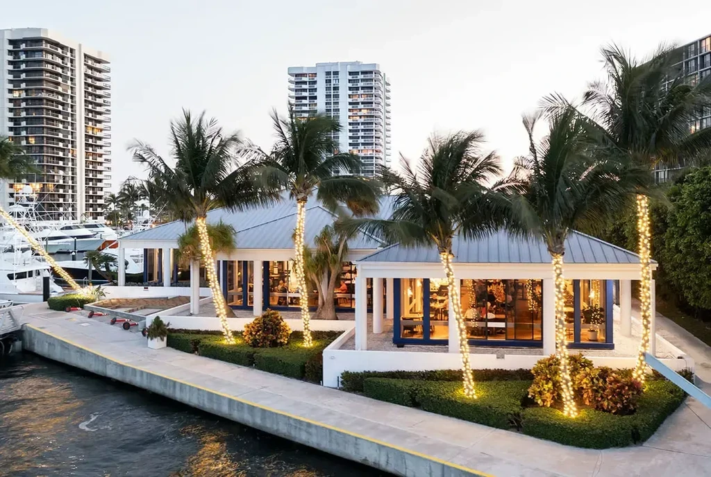 Waterfront restaurant with lit palm trees, yachts, and city skyline.