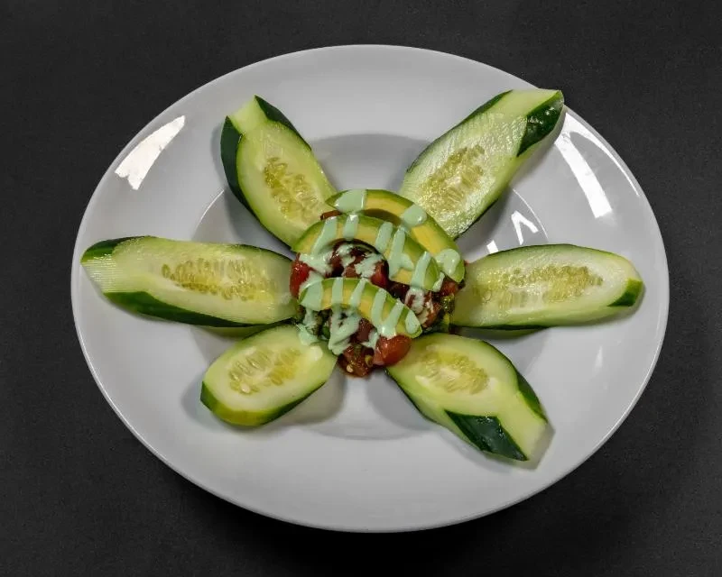 Elegant dish with radial cucumber slices, red food mound, avocado, green sauce.