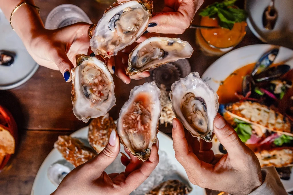 Multiple hands hold fresh oysters with sauce at a dining table.