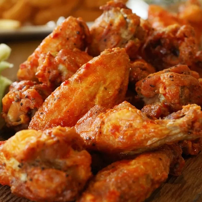 Pile of buffalo chicken wings with reddish-orange sauce, blurred fries and celery.