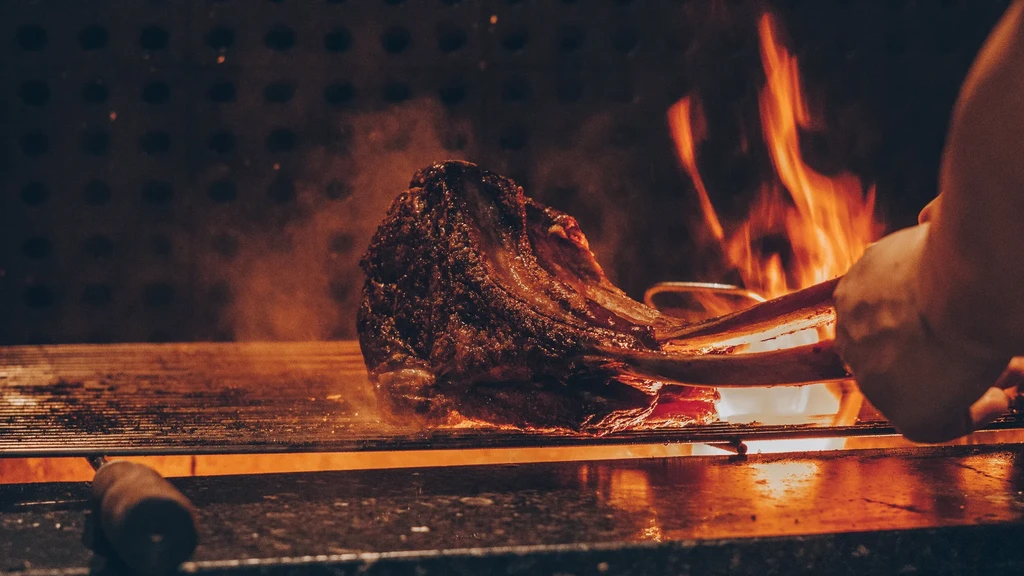 Tomahawk steak searing over intense open flames, held by tongs.