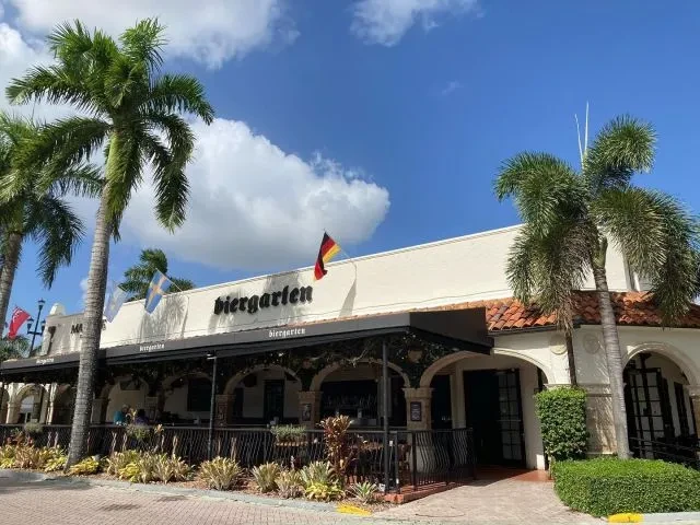 Biergarten restaurant exterior with red roof, dark awning, flags, palm trees.