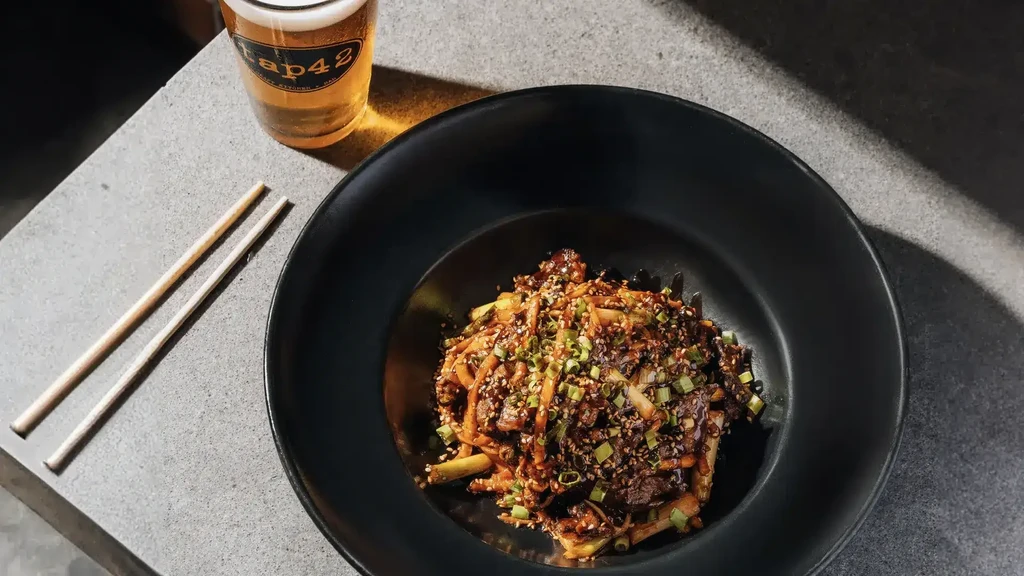 Overhead of stir-fry in black bowl with chopsticks and Tap42 beer.