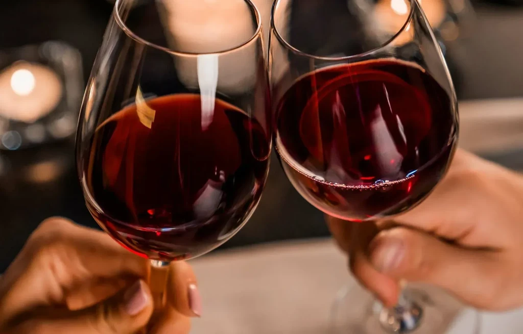 Two hands clinking red wine glasses against soft blurred lights.