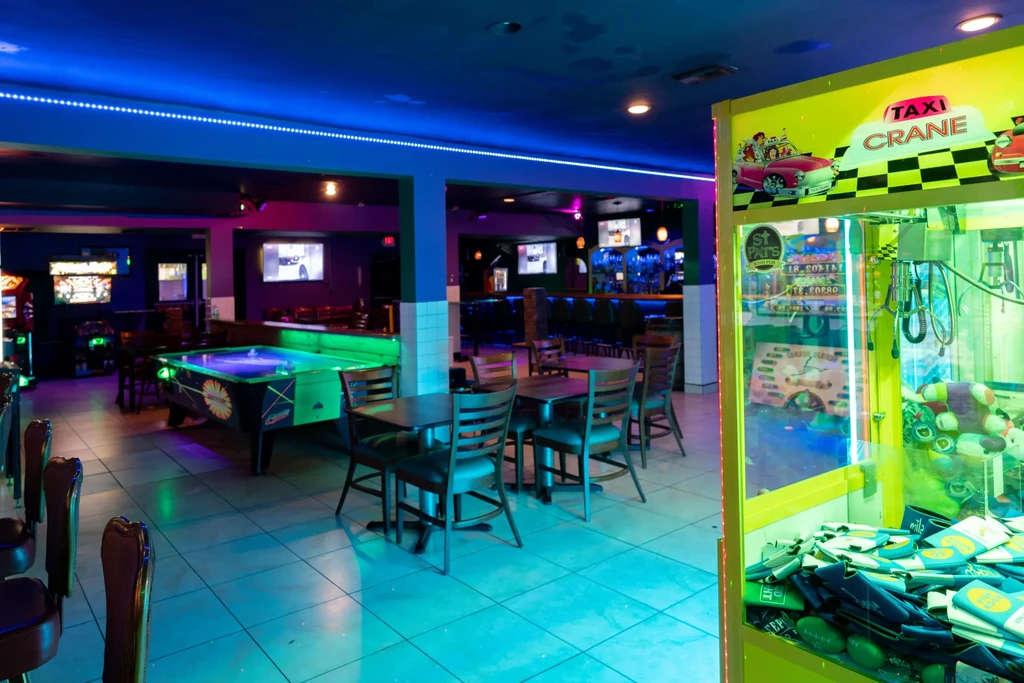 Neon-lit arcade/bar interior with claw machine, pool table, arcade games.