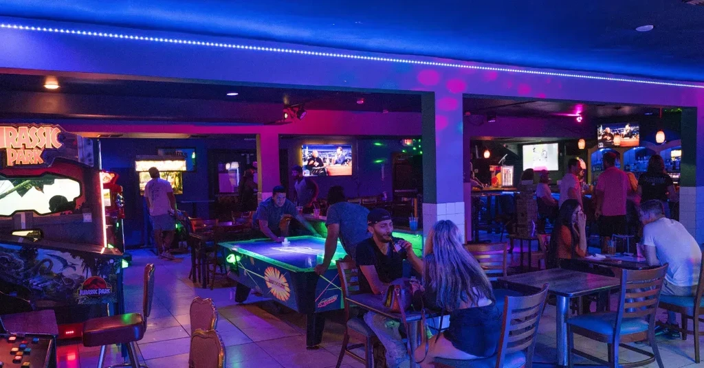 Neon-lit bar interior with patrons playing arcade and air hockey.