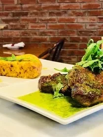 White plate with seared meat, arugula, green sauce, and yellow polenta.