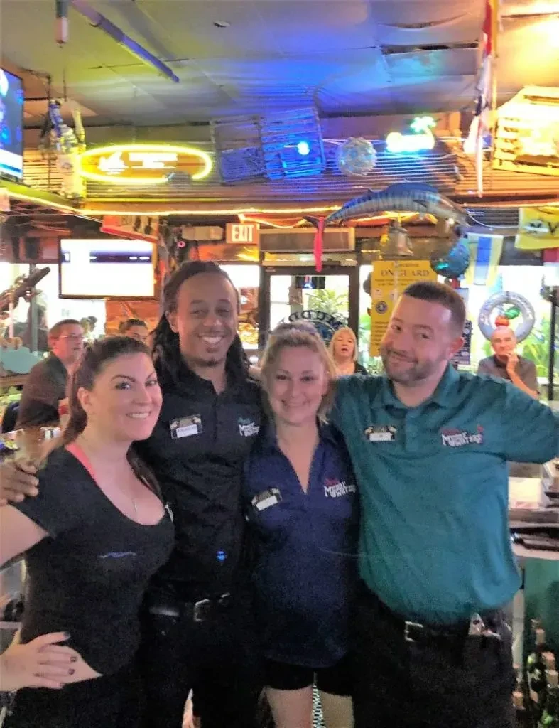 Four uniformed staff smile in a vibrant bar with neon lights.