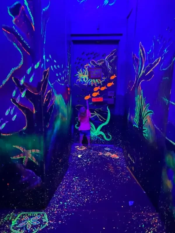 Child in blacklight hallway with glowing neon underwater murals.