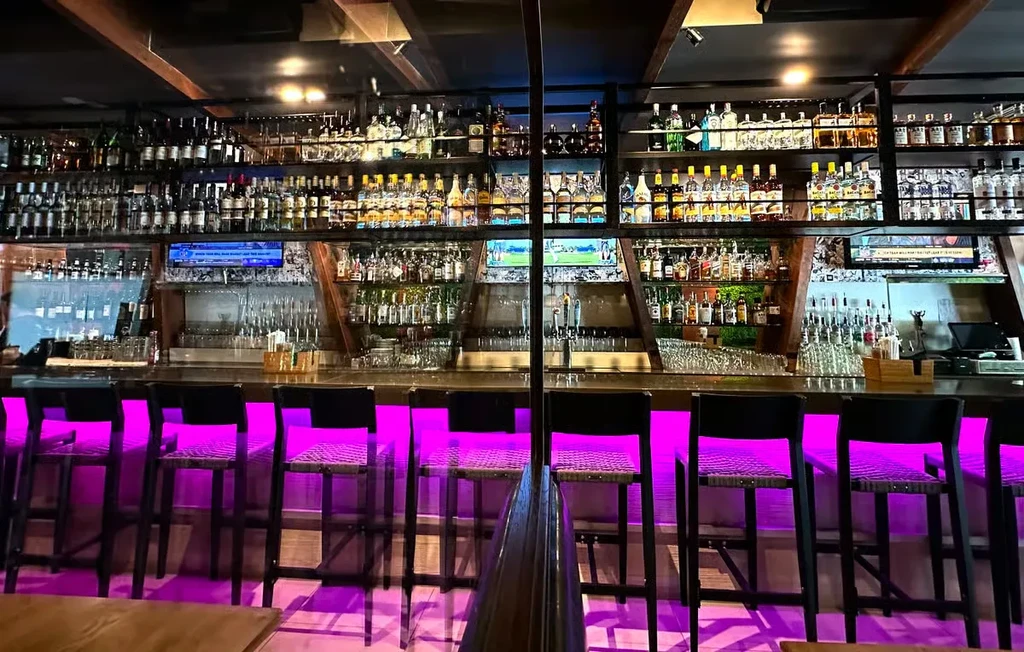 Modern bar with purple LED lighting, bar stools, and liquor bottles.
