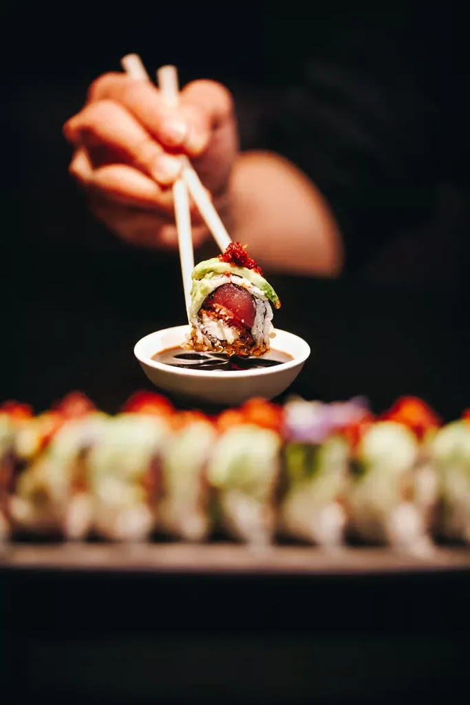 Chopsticks hold a tuna, cream cheese, and avocado sushi roll over soy sauce.