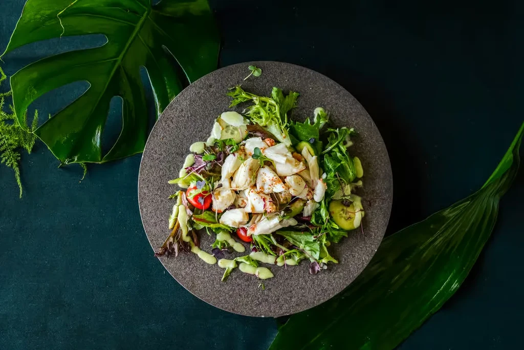 Plated crab and vegetable salad with dressing, framed by tropical leaves.